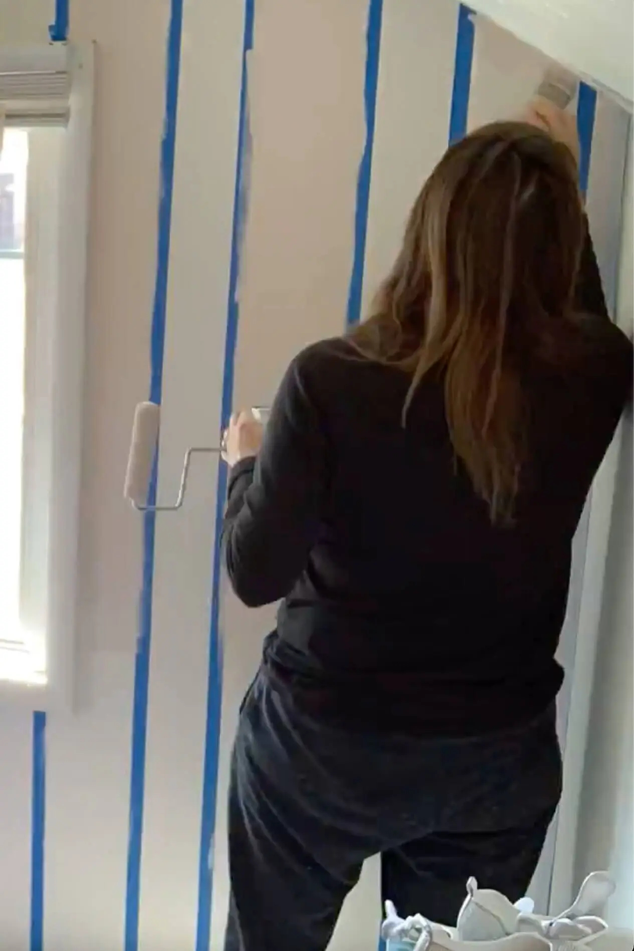 Woman painting vertical stripes with a roller on a bedroom wall, starting the buffalo plaid pattern for a DIY accent wall project